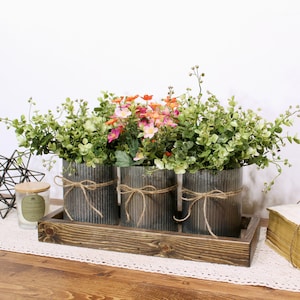 May include: Three galvanized metal planters with rustic twine bows, filled with green leafy plants and pink and orange flowers, sitting on a wooden tray.
