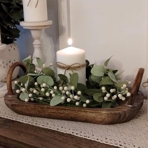 May include: A wooden tray with handles, filled with white berries and green leaves, and three white pillar candles. Two of the candles are on white pedestals.
