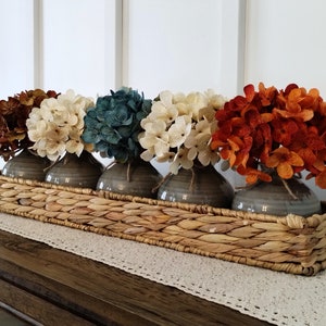 May include: Five faux hydrangea flower arrangements in grey ceramic vases, arranged in a woven wicker basket. The flowers are in shades of brown, cream, blue, and orange.