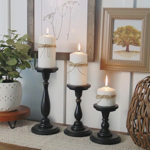 May include: Three black candle holders with white candles on a wooden table with a white lace runner. The candles are tied with twine and have a rustic look.