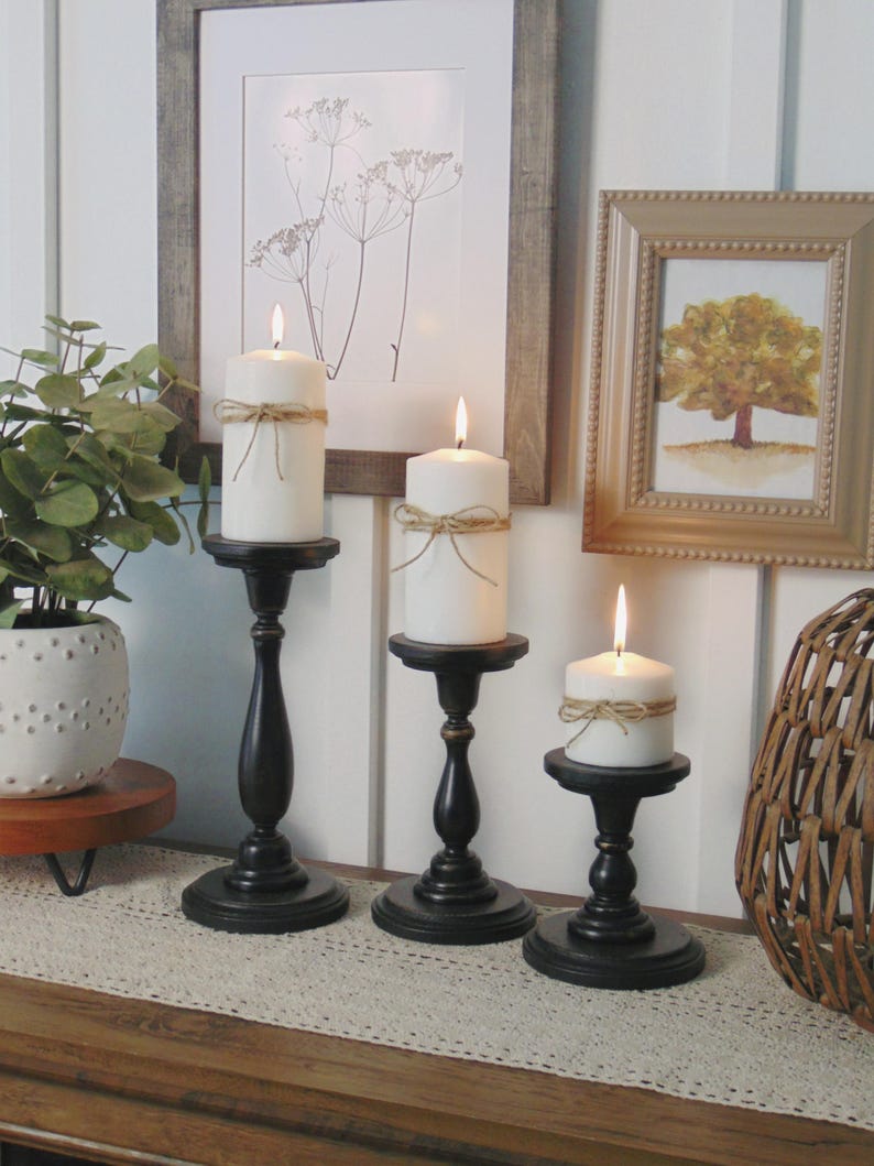 May include: Three black candle holders with white candles on a wooden table with a white lace runner. The candles are tied with twine and have a rustic look.