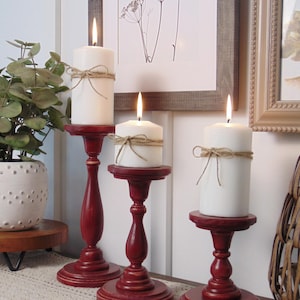 May include: Three red wooden candle holders with white candles. The candles are tied with twine. The candle holders are on a beige table runner.