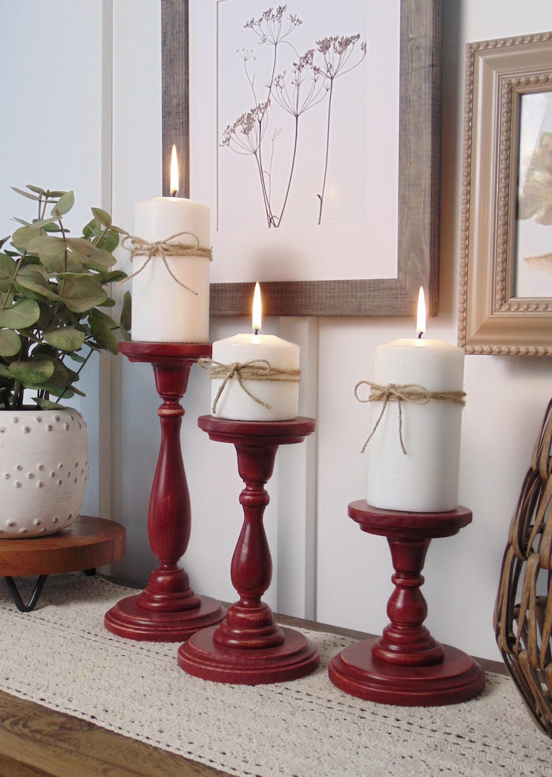May include: Three red wooden candle holders with white candles. The candles are tied with twine. The candle holders are on a beige table runner.