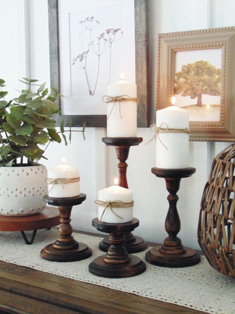 May include: Four white pillar candles with twine accents on wooden candle holders. The candles are lit and the holders are dark brown wood. The candles are on a table with a white lace runner and a brown wooden surface.