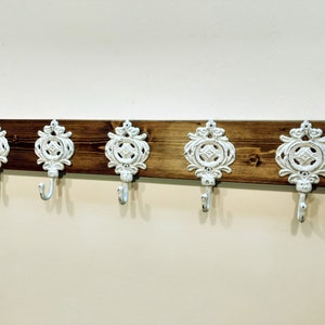 May include: A wooden wall-mounted coat rack with five white metal hooks. The hooks are ornate and have a decorative design. The wood is a dark brown colour.