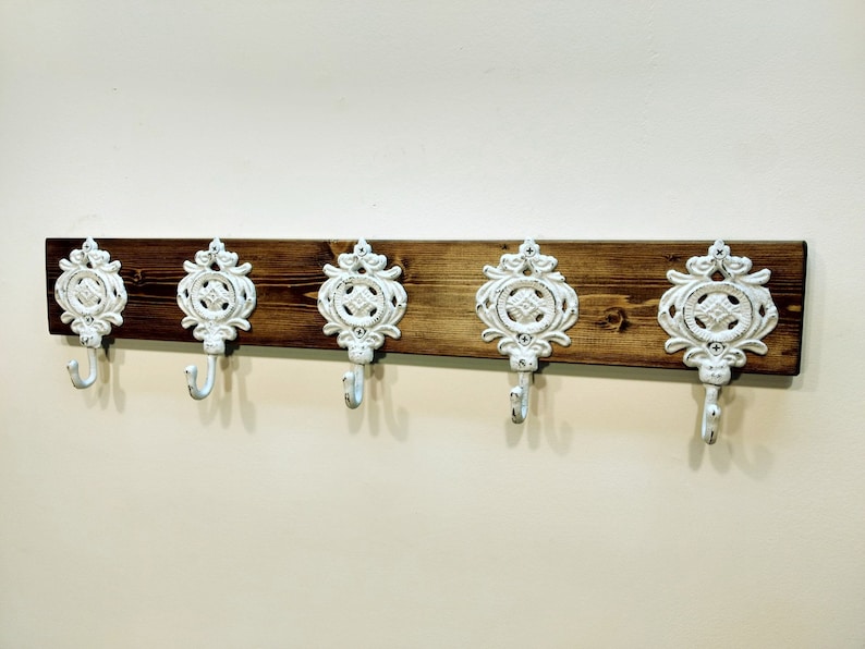May include: A wooden wall-mounted coat rack with five white metal hooks. The hooks are ornate and have a decorative design. The wood is a dark brown colour.