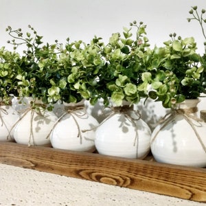 May include: Five white ceramic vases with a rustic twine accent, filled with green leafy sprigs, arranged in a wooden tray.