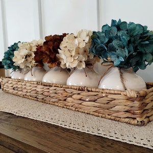 May include: A woven wicker tray with four white ceramic vases filled with faux hydrangea flowers in shades of teal, cream, and brown.