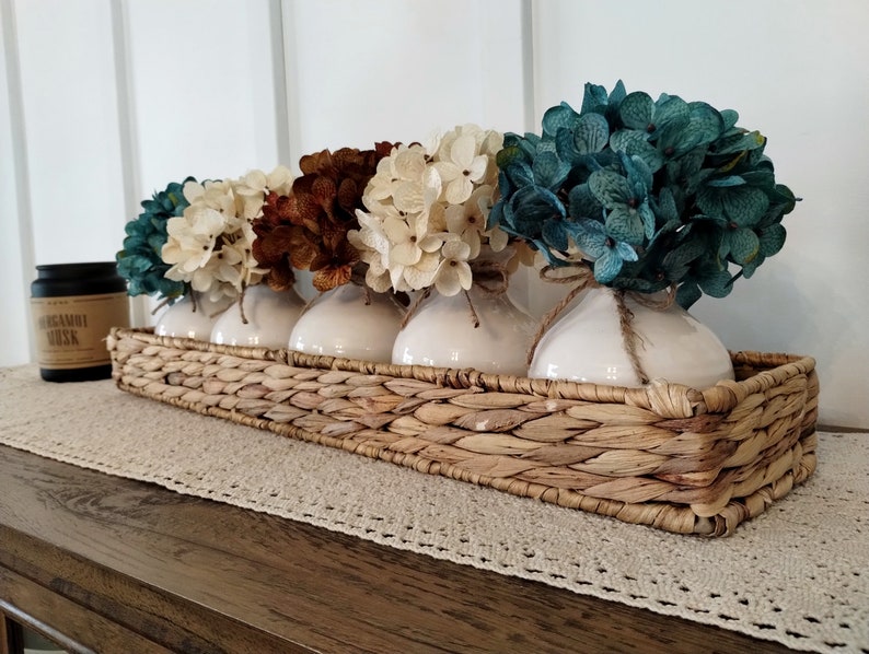 May include: A woven wicker tray with four white ceramic vases filled with faux hydrangea flowers in shades of teal, cream, and brown.