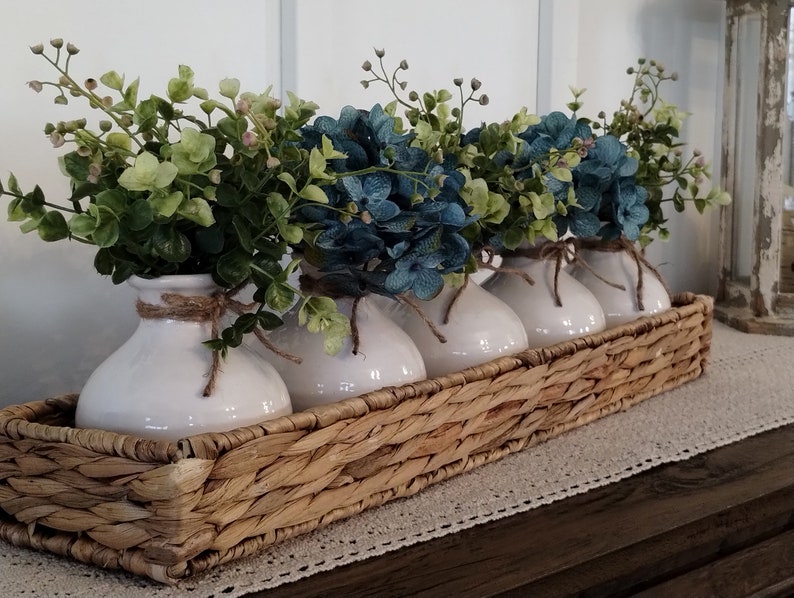 May include: A woven wicker basket filled with four white ceramic vases, each holding a bouquet of blue and green faux flowers.