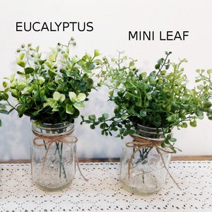 May include: Two mason jars filled with green eucalyptus and mini leaf greenery. The jars are tied with twine and sit on a white lace tablecloth.