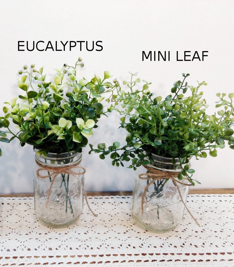 May include: Two mason jars filled with green eucalyptus and mini leaf greenery. The jars are tied with twine and sit on a white lace tablecloth.