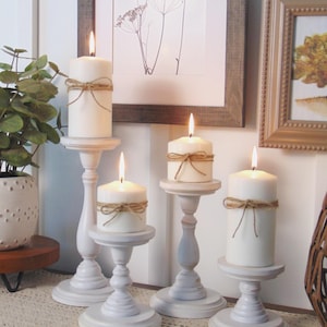 May include: Set of four white wooden candle holders with a distressed finish. Each holder features a simple design with a round base and a tall, slender column. The candles are white and have rustic twine tied around them.