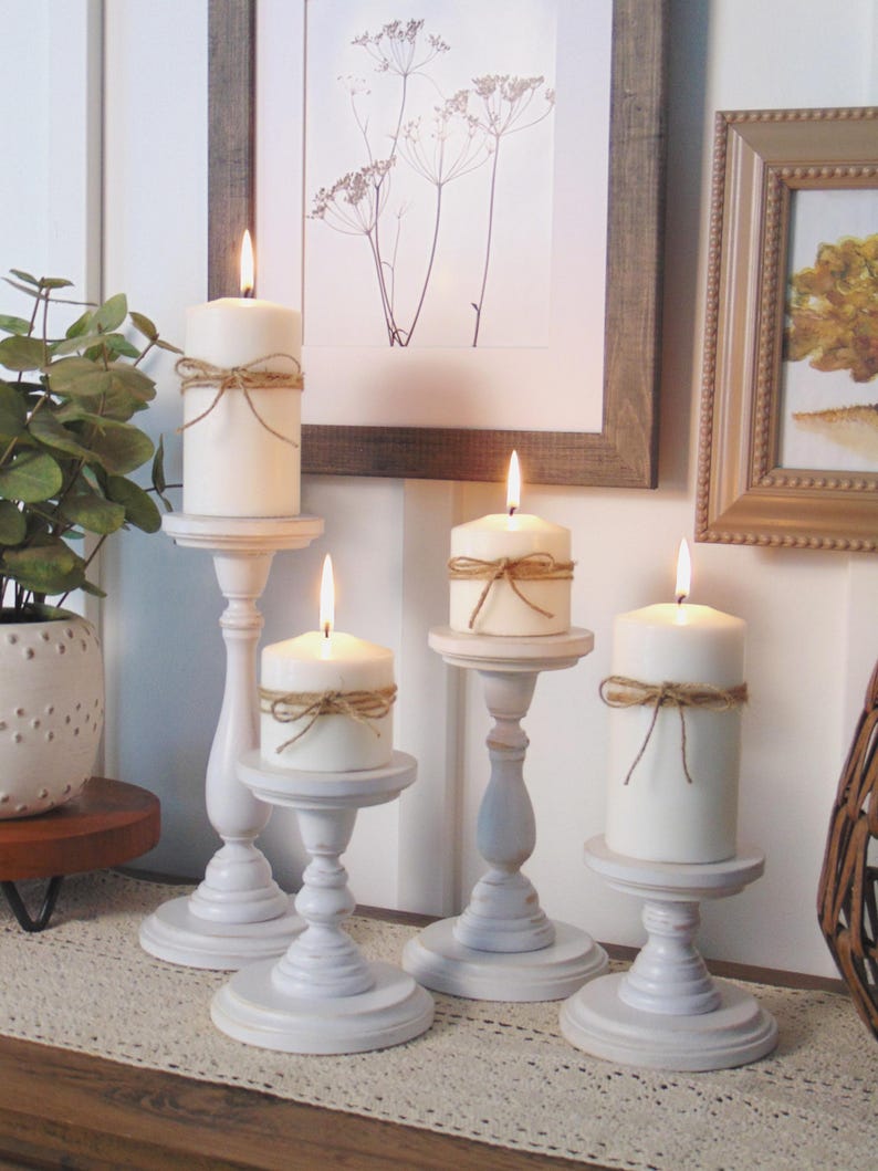 May include: Set of four white wooden candle holders with a distressed finish. Each holder features a simple design with a round base and a tall, slender column. The candles are white and have rustic twine tied around them.