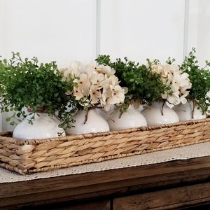 May include: A woven wicker basket filled with five white ceramic vases, each holding a small bouquet of white hydrangeas and green foliage.
