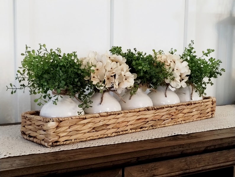 May include: A woven wicker basket filled with five white ceramic vases, each holding a small bouquet of white hydrangeas and green foliage.