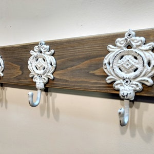 May include: A rustic wooden wall-mounted coat rack with three white cast iron hooks. The hooks have a decorative design with a circular centre and intricate scrollwork.