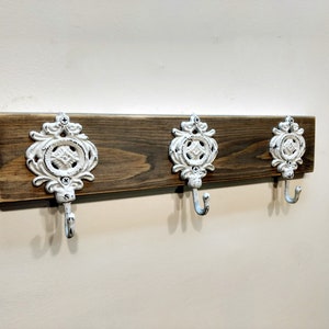 May include: A rustic wooden wall mounted coat rack with three white metal hooks. The hooks have a decorative design.