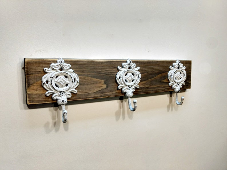 May include: A rustic wooden wall mounted coat rack with three white metal hooks. The hooks have a decorative design.
