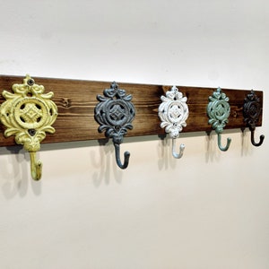 May include: A wooden wall-mounted coat rack with five decorative hooks. The hooks are cast iron and painted in various colours, including yellow, grey, white, green, and brown.