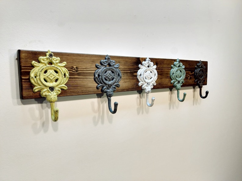May include: A wooden wall-mounted coat rack with five decorative hooks. The hooks are cast iron and painted in various colours, including yellow, grey, white, green, and brown.