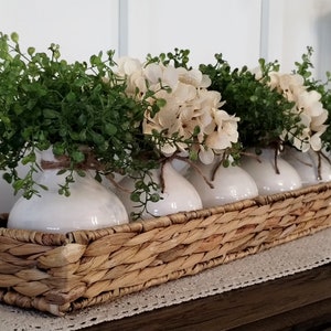May include: A woven wicker tray with five white ceramic vases filled with green leafy sprigs and cream-colored flowers.