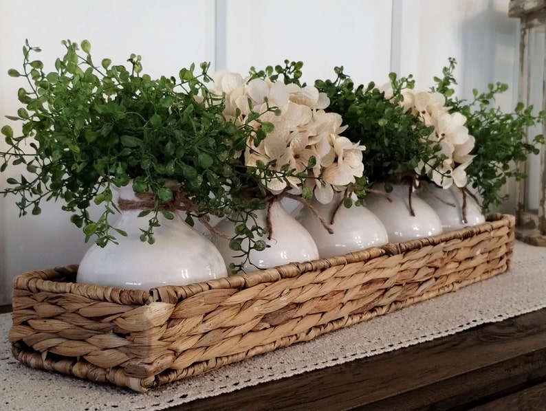 May include: A woven wicker tray with five white ceramic vases filled with green leafy sprigs and cream-colored flowers.