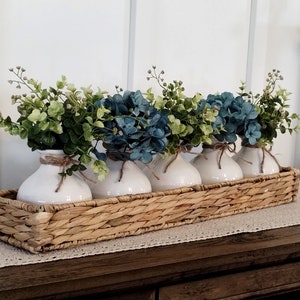 May include: Five white ceramic vases with blue and green floral arrangements are displayed in a woven wicker basket. The basket is on a wooden surface with a white lace runner.