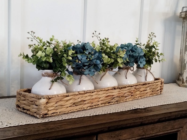 May include: Five white ceramic vases with blue and green floral arrangements are displayed in a woven wicker basket. The basket is on a wooden surface with a white lace runner.