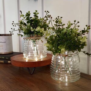 May include: Two clear glass jars with a textured surface, filled with green leafy plants and white fairy lights. The jars are sitting on a wooden stand with black metal legs.
