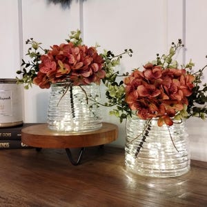 May include: Two clear glass vases with textured, horizontal lines, each filled with orange-red flowers and greenery. The vases sit on a wooden stand with a black metal base. A candle and books are in the background.