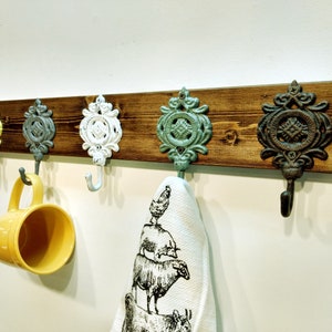 May include: A rustic wooden wall-mounted rack with five decorative metal hooks. The hooks are painted in various colours, including yellow, blue, green, and white. A yellow mug hangs from one of the hooks, and a white tea towel hangs from another.