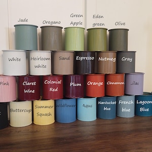 May include: A collection of 24 small, cylindrical planters in various colours, including black, white, pink, red, orange, green, blue, and grey. Each planter has a ribbed texture and is labelled with a different colour name.