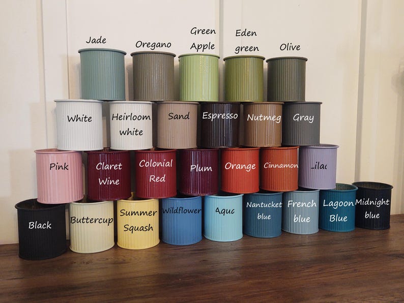 May include: A collection of 24 small, cylindrical planters in various colours, including black, white, pink, red, orange, green, blue, and grey. Each planter has a ribbed texture and is labelled with a different colour name.
