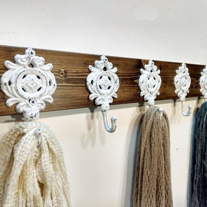 May include: A wooden wall-mounted coat rack with four white ornate hooks. The hooks have a decorative design and are attached to a dark brown wooden board. Three items are hanging from the hooks.