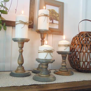 May include: Five wooden candle holders, painted in shades of grey and brown, each holding a white pillar candle with twine wrapped around the base. The candles are lit and the holders are arranged on a white table runner.