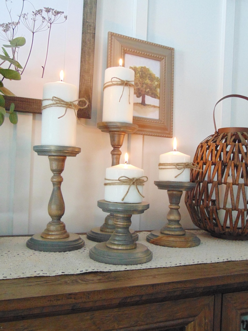 May include: Five wooden candle holders, painted in shades of grey and brown, each holding a white pillar candle with twine wrapped around the base. The candles are lit and the holders are arranged on a white table runner.