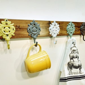 May include: A rustic wooden wall mounted coat rack with five decorative metal hooks. The hooks are painted in various colours, including yellow, white, blue, and green. A yellow mug hangs from one hook and a white kitchen towel with a black and white sheep design hangs from another hook.