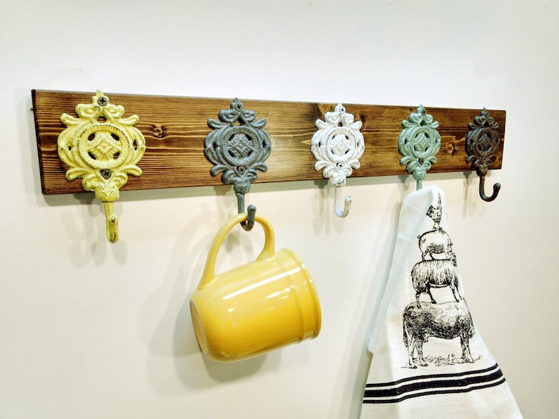 May include: A rustic wooden wall mounted coat rack with five decorative metal hooks. The hooks are painted in various colours, including yellow, white, blue, and green. A yellow mug hangs from one hook and a white kitchen towel with a black and white sheep design hangs from another hook.