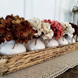 May include: A wicker basket filled with five white ceramic vases, each holding a different coloured bouquet of faux hydrangeas. The flowers are shades of brown, white, red, and burgundy.