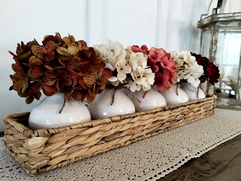 May include: A wicker basket filled with five white ceramic vases, each holding a different coloured bouquet of faux hydrangeas. The flowers are shades of brown, white, red, and burgundy.