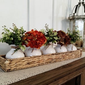 May include: Four white ceramic vases with orange and brown flowers arranged in a woven wicker basket. The basket is on a wooden table with a white crocheted runner.