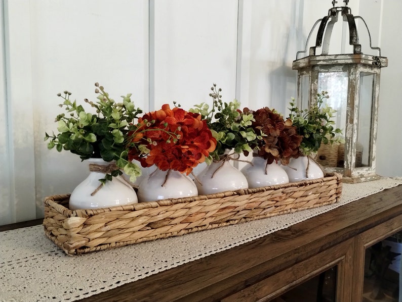 May include: Four white ceramic vases with orange and brown flowers arranged in a woven wicker basket. The basket is on a wooden table with a white crocheted runner.