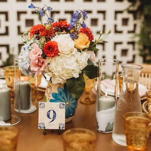May include: A table number nine with the word "Nueve" written below it. The table number is on a blue and white tile with a floral design. The tile is on a black stand. A bouquet of flowers in a blue and white floral vase is in the background. The flowers are red, white, yellow, and blue.