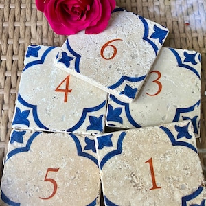 May include: Five ceramic tile coasters with blue and white floral designs and orange numbers 1 through 6. The coasters are arranged in a semi-circle on a woven wicker surface.