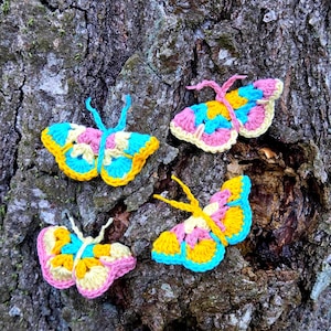 May include: Four colorful crochet butterflies in yellow, pink, and blue are displayed against a textured tree bark background. Each butterfly has a unique pattern and shape, showcasing handmade craftsmanship. These decorative butterflies are perfect for adding a whimsical touch to any space.