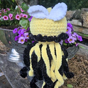 May include: A crocheted plush toy shaped like a bee with a round yellow head, two white wings, and a body featuring alternating long, dangling black and yellow yarn tentacles. The plush has a textured, soft appearance with a black collar detail.