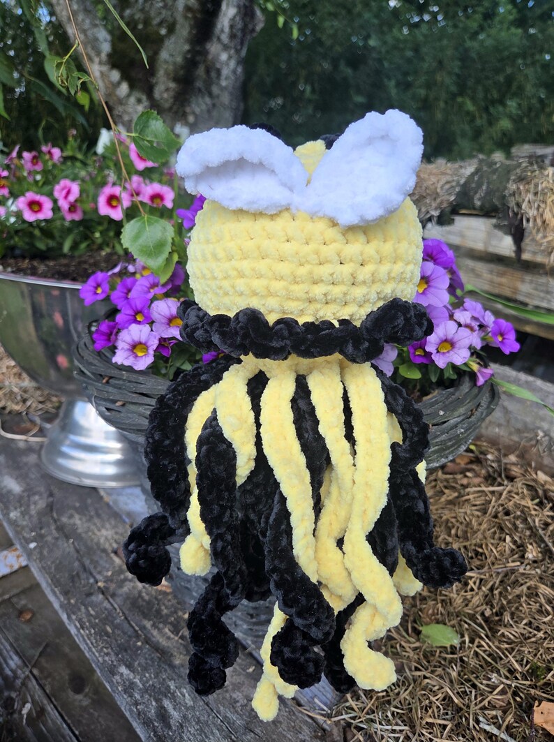 May include: A crocheted plush toy shaped like a bee with a round yellow head, two white wings, and a body featuring alternating long, dangling black and yellow yarn tentacles. The plush has a textured, soft appearance with a black collar detail.
