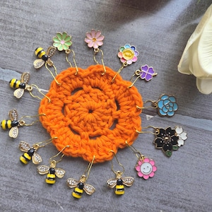 Könnte beinhalten: Eine Auswahl an farbenfrohen Maschenmarkierern, darunter Bienen- und Blumenmotive, die um einen orangefarbenen gehäkelten Kreis angeordnet sind. Die Maschenmarkierer haben goldfarbene Metallbeschläge. Die Blumen sind in verschiedenen Farben, darunter Rosa, Blau und Lila.