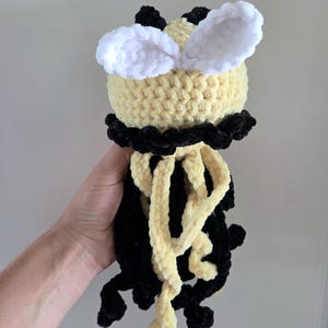May include: A handmade crochet plush toy featuring a round yellow head with two white fuzzy ears, a black collar, and dangling black and yellow yarn tentacles. The soft, textured yarn construction creates a tactile surface with distinct color blocks.